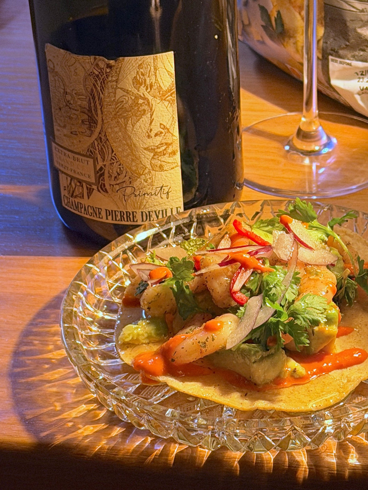 Bottle of Champagne Pierre Deville Primitif beside a prawn taco on a cut glass plate